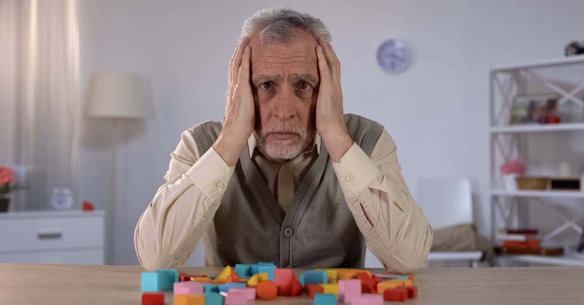 an elderly person with hands on head looking frustrated over colorful building blocks, suggesting challenges with cognitive tasks.