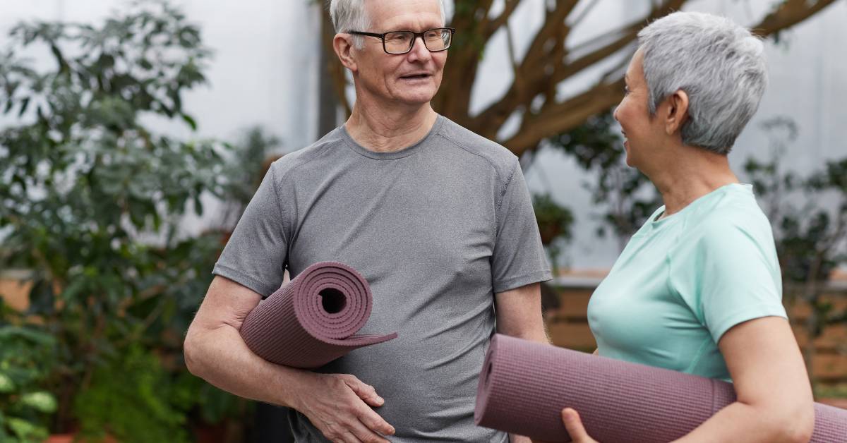 Jóga pro seniory (Youtube): Zacvičte si v pohodlí domova 1 senior couple holding yoga mats outdoors, smiling and preparing for a relaxed fitness session focused on flexibility and well-being.