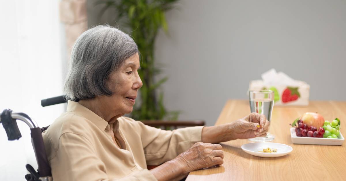 Léky na paměť pro seniory: Seznam nejúčinnějších léků a přírodních doplňků stravy 5 an elderly person in a wheelchair taking small pills, possibly naturální doplňky stravy, at a table with fresh fruit and water.