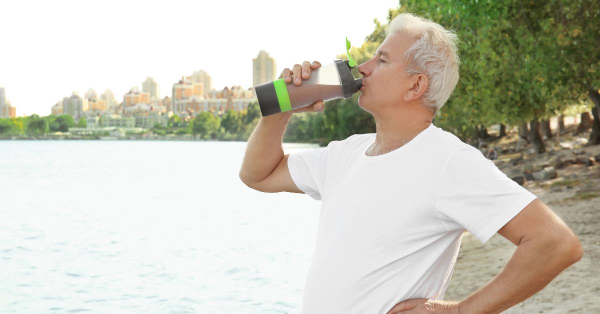 4 Nejlepší Nutridrinky pro seniory: Co to je a jaké jsou jeho výhody? 2 an older adult drinking from a nutridrink bottle by a scenic waterfront during outdoor exercise.