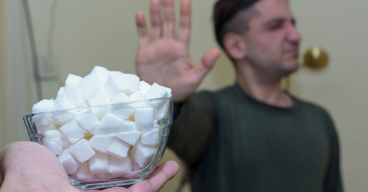 8 Důvodů Proč Nejíst Cukr & Jak Omezit Cukr A Čím Ho Zdravě Nahradit 3 a hand holding a bowl of cubed white sugar while raising the other hand to stop, emphasizing excess sugar intake