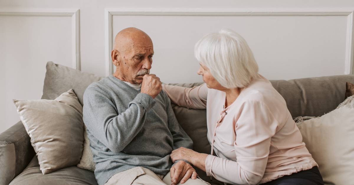 Babské rady na kašel: 18 Nejlepších tipů jak se zbavit kašle přírodní cestou bez léků! 1 an elderly couple sitting closely on a couch, one comforting the other with a supportive hand gesture during what appears to be a moment of concern or discussion.