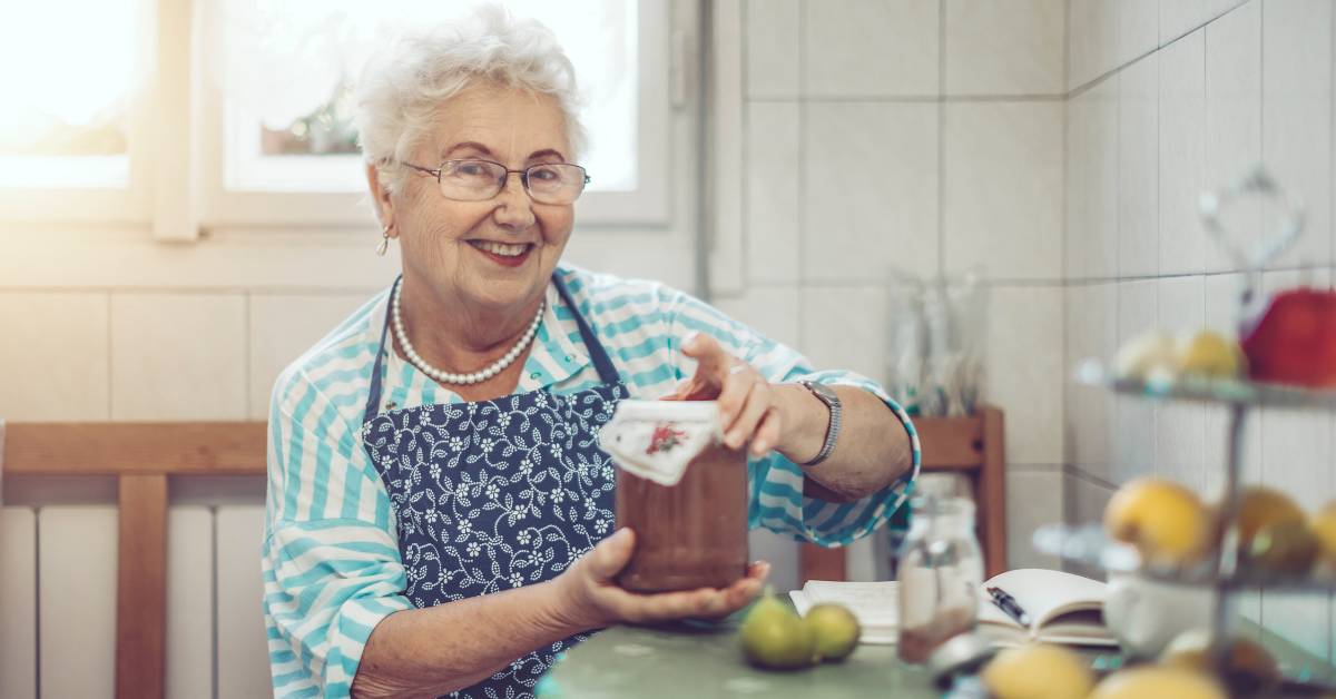 Babské rady na plíseň nehtů: 20 Nejlepších tipů jak se jí zbavit doma přírodní cestou bez léků 1 a smiling elderly woman wearing an apron holding a wooden spoon with a recipe book open, preparing natural home remedies in a bright kitchen.