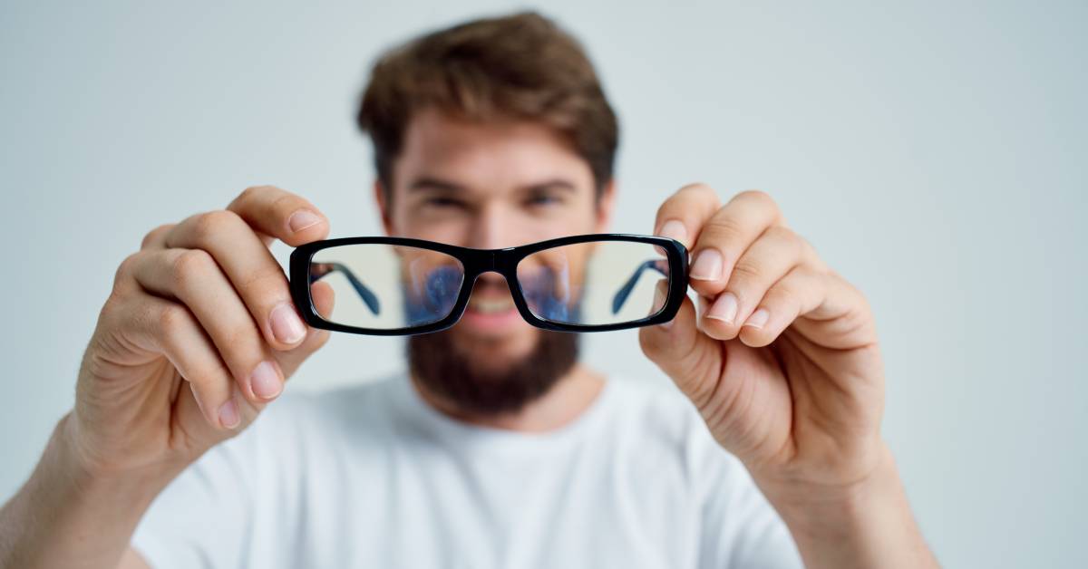 Batesova Metoda Zlepšení Zraku: Jak ji používat a co můžete očekávat 4 a person holding up a pair of black-rimmed glasses in front of their face, demonstrating visual focus or eye exercise.