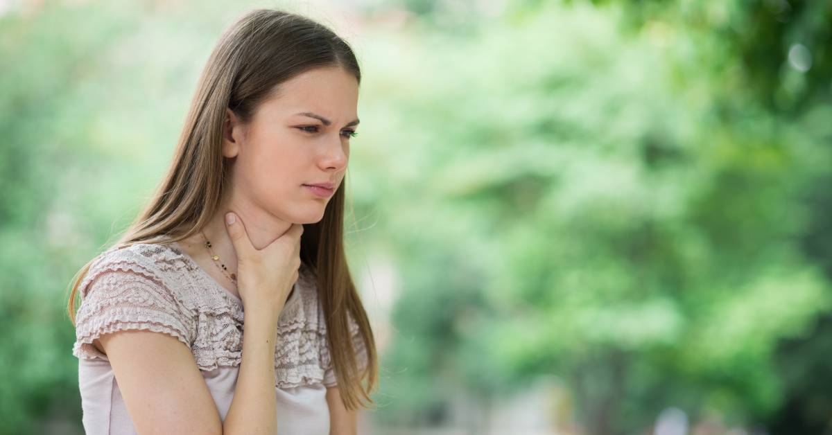 Babské rady na bolest v krku: 21 Nejlepších způsobů přírodní léčby! 1 a woman holding her throat, appearing to experience discomfort or bolest v krku outdoors.
