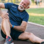 Senior Hub - Svět Pomůcek Pro Život Bez Bolesti A Limitací! 34 an elderly man sitting on a track while holding and stretching his painful knee, showing signs of discomfort or injury related to knee strain or overuse.