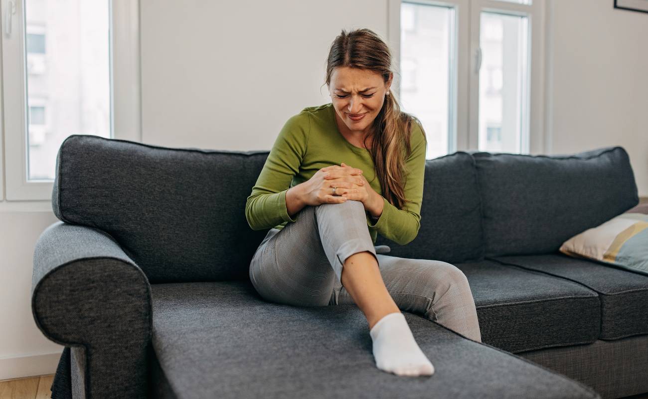 a person sitting on a couch holding and bending their painful knee, showing signs of discomfort from knee joint pain.